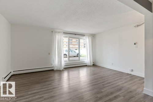 Spacious room featuring wood-finish flooring, white walls, and an expansive window with white drapes - 101 8604 Gateway Boulevard, Edmonton, AB - Indoor