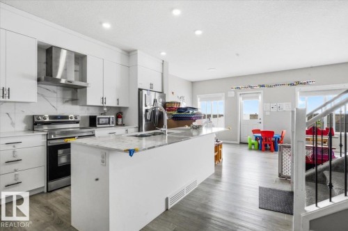 Modern kitchen featuring white cabinetry, a large center island with a sink, stainless steel appliances, marble-look backsplash, and wood-finish flooring - 7056 182 Avenue, Edmonton, AB - Indoor Photo Showing Kitchen With Upgraded Kitchen