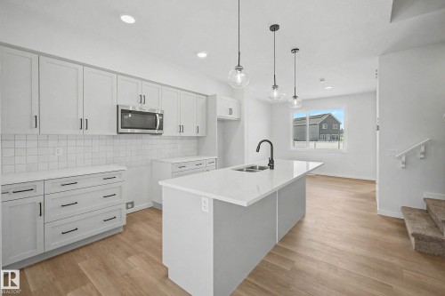 Modern kitchen featuring a central island with a double basin sink, light wood-finish flooring, white cabinetry with black hardware, a stainless steel microwave, and pendant lighting - 720 Southfork Drive, Leduc, AB - Indoor Photo Showing Kitchen With Double Sink With Upgraded Kitchen
