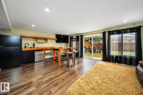 Spacious interior featuring wood-finish flooring, recessed lighting, and a built-in wet bar with a black refrigerator and stainless steel under-counter appliance - 126 Harrison Drive, Edmonton, AB - Indoor