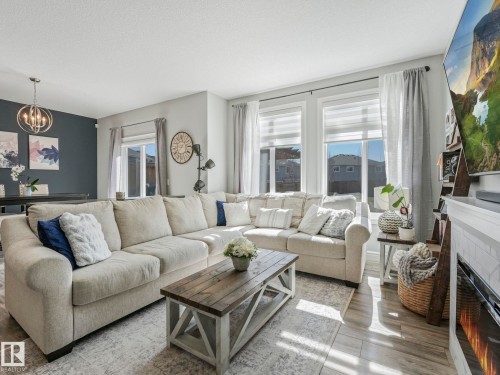 Open-concept living area featuring light wood-finish flooring, multiple windows with blinds, a white fireplace surround with a tile accent, and a built-in electric fireplace - 3817 Robins Crescent, Edmonton, AB - Indoor Photo Showing Living Room With Fireplace