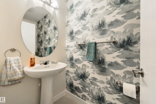 Pedestal sink with chrome faucet, oval mirror, wall-mounted lighting, and an accent wall featuring patterned wallpaper - 4914 47 Street, Redwater, AB - Indoor Photo Showing Bathroom