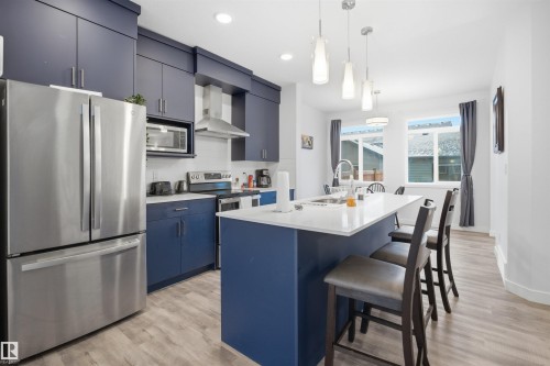 The kitchen features rich blue cabinetry, stainless steel appliances, and an island with a white countertop and seating for three - 4047 Hawthorn Link Link, Edmonton, AB - Indoor Photo Showing Kitchen With Upgraded Kitchen