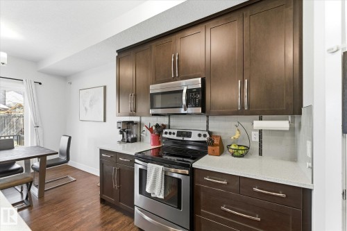Modern kitchen featuring dark wood-finish cabinetry with stainless hardware, light-toned countertops, and a subway tile backsplash - 54 Cypress Link, Fort Saskatchewan, AB - Indoor Photo Showing Kitchen With Upgraded Kitchen