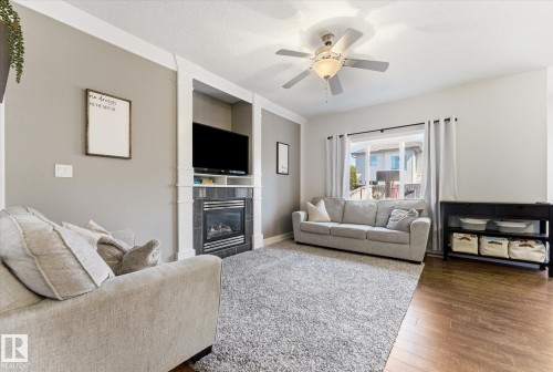 Living area featuring a built-in fireplace with dark gray tile surround and white mantel, a ceiling fan with integrated lighting, wood-finish flooring, and a large window providing natural light - 54 Cypress Link, Fort Saskatchewan, AB - Indoor Photo Showing Living Room With Fireplace