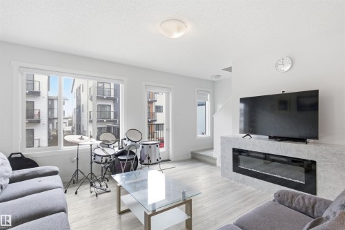 Living area featuring wood-finish flooring, a wall-mounted electric fireplace with a stone-look surround, and abundant natural light from large windows and a glass-paneled door - 9 850 Secord Boulevard, Edmonton, AB - Indoor Photo Showing Living Room With Fireplace