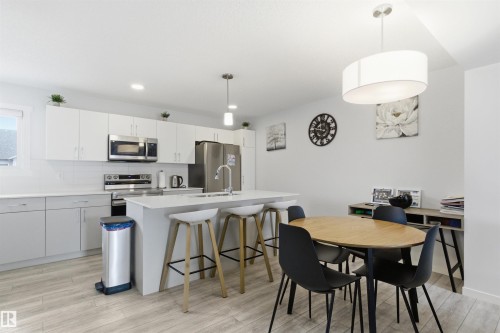 Open-concept kitchen and dining area featuring light wood-finish flooring, white cabinetry, stainless steel appliances, a center island with bar seating, and a modern drum pendant light fixture - 9 850 Secord Boulevard, Edmonton, AB - Indoor