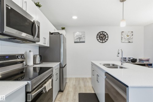 Modern kitchen featuring a central island with double basin sink, stainless steel appliances, white cabinetry, wood-finish flooring, and recessed lighting - 9 850 Secord Boulevard, Edmonton, AB - Indoor Photo Showing Kitchen With Double Sink