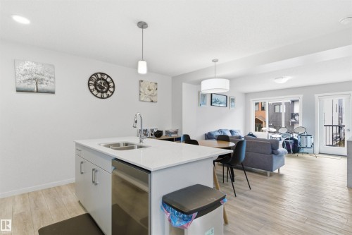 Open-concept living space featuring a kitchen island with dual basin sink and stainless steel dishwasher, wood-finish flooring, recessed lighting, and a sliding glass door leading to a balcony - 9 850 Secord Boulevard, Edmonton, AB - Indoor
