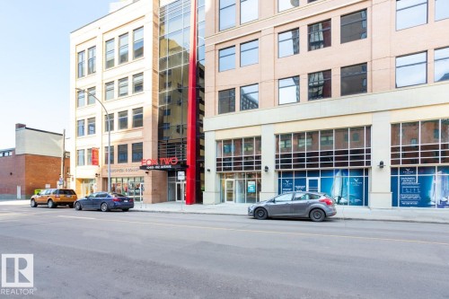 Modern commercial building featuring a tan brick and stone exterior, a prominent red architectural accent, expansive ground-floor retail windows, and a glass-enclosed vertical element - #1603 10410 102 Av Nw, Edmonton, AB - Outdoor