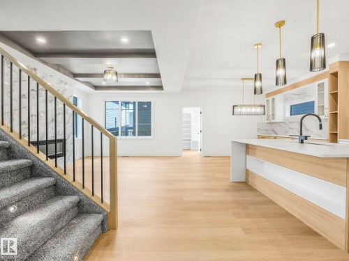 Open-concept living area featuring light wood-finish flooring, a contemporary kitchen island with a white countertop and integrated sink, and an elegant staircase with carpeted treads and an open black metal railing - 6125 Crawford Drive, Edmonton, AB - Indoor