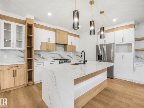 Modern kitchen featuring a waterfall island with a double basin sink, light wood-finish flooring, white and wood-finish cabinetry, stainless steel appliances, and a marble-look backsplash - 6125 Crawford Drive, Edmonton, AB - Indoor Photo Showing Kitchen With Double Sink With Upgraded Kitchen