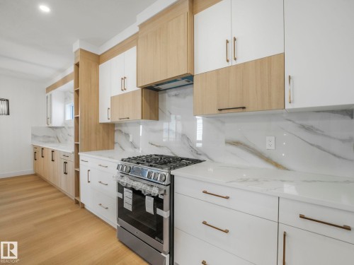 Kitchen featuring a stainless steel gas range, two-tone cabinetry with white and light wood-finish elements, white countertops with veining, a matching full-height backsplash, and wood-finish flooring - 6125 Crawford Drive, Edmonton, AB - Indoor Photo Showing Kitchen