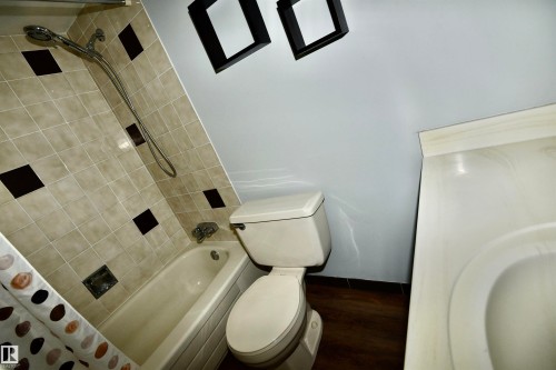 Bathroom featuring a bathtub with tiled surround, a toilet, a vanity with an integrated sink, and wood-finish flooring - 21 10160 119 Street Nw, Edmonton, AB 
