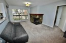Living room featuring a stone-clad fireplace with a wood mantel, light gray carpeting, and a sliding glass door leading to a balcony - 21 10160 119 Street Nw, Edmonton, AB 