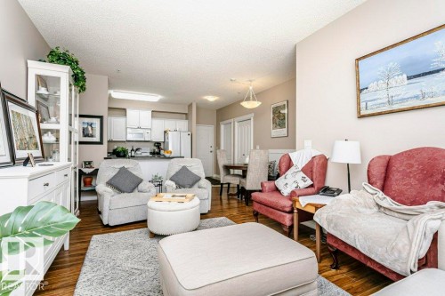 Open-concept living area with wood-finish flooring, neutral wall tones, and a kitchen featuring white cabinetry, a double-basin sink, and stainless steel appliances - 323 16807 100 Avenue, Edmonton, AB - Indoor Photo Showing Living Room