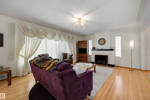 Living area featuring wood-finish flooring, a decorative fireplace with a dark mantel, glass block windows, and a multi-tiered ceiling light fixture - 9602 85 Street, Edmonton, AB - Indoor Photo Showing Living Room With Fireplace