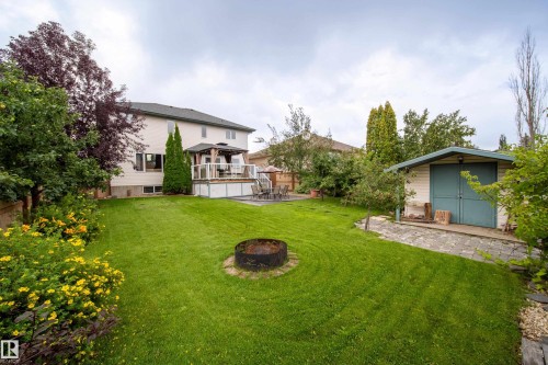 The property features an expansive lawn with a fire pit, a shed with blue double doors, and a stone path - 4 Windrose Drive, Leduc, AB - Outdoor With Deck Patio Veranda With Backyard With Exterior