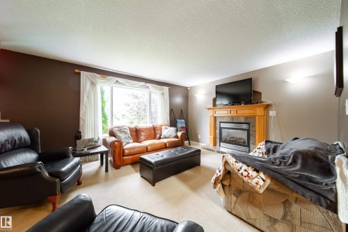 The living area features a large window with white curtains, carpeted flooring, and a fireplace with a light wood mantel and stone surround - 4 Windrose Drive, Leduc, AB - Indoor Photo Showing Living Room With Fireplace