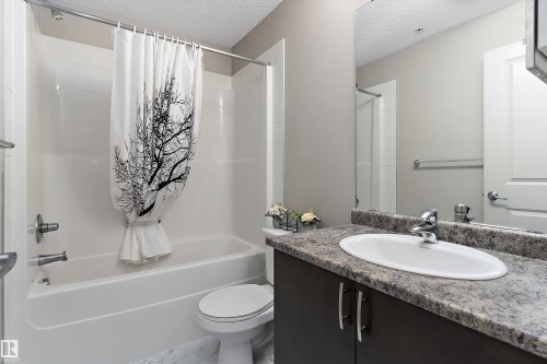 Bathroom featuring a white bathtub and shower combination, a dark wood-finish vanity with a speckled countertop, an oval undermount sink, and a large wall-mounted mirror - 221 344 Windermere Road, Edmonton, AB - Indoor Photo Showing Bathroom