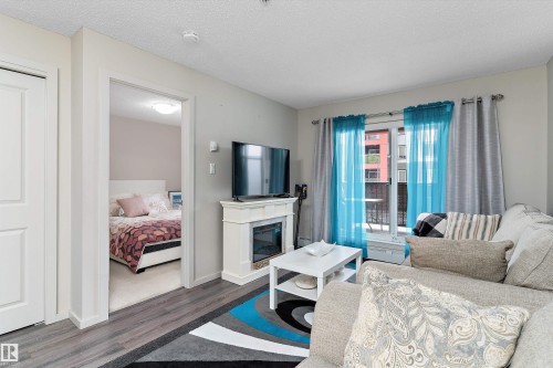 Living area featuring wood-finish flooring, a built-in electric fireplace with mantel, and a balcony with sliding glass doors - 221 344 Windermere Road, Edmonton, AB - Indoor Photo Showing Other Room With Fireplace