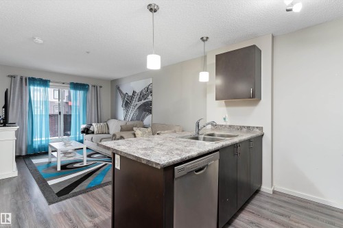 Open-concept living area featuring wood-finish flooring, a kitchen island with a double basin sink, stainless steel dishwasher, and dark cabinetry with a coordinating countertop - 221 344 Windermere Road, Edmonton, AB - Indoor Photo Showing Kitchen With Double Sink