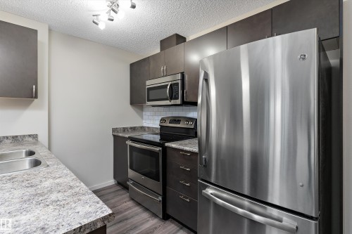 Contemporary kitchen featuring dark wood-finish cabinetry, stainless steel appliances, a subway tile backsplash, and wood-finish flooring - 221 344 Windermere Road, Edmonton, AB - Indoor Photo Showing Kitchen With Double Sink