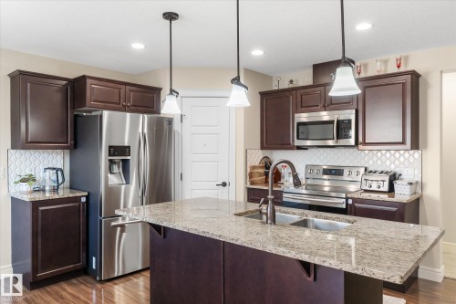 Contemporary kitchen featuring dark wood cabinetry, stainless steel appliances, granite countertops, a diamond-patterned backsplash, and pendant lighting over a dual-basin sink - 6864 Evans Wynd, Edmonton, AB - Indoor Photo Showing Kitchen With Stainless Steel Kitchen With Double Sink With Upgraded Kitchen