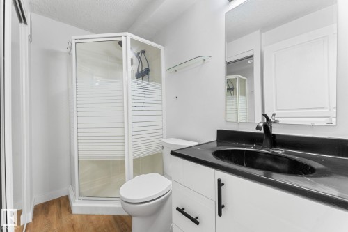 Compact bathroom featuring a corner shower with glass enclosure, white vanity with black countertop and integrated sink, matte black faucet, and wood-finish flooring - 362 Blackburn Drive E, Edmonton, AB - Indoor Photo Showing Bathroom