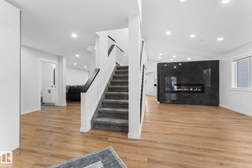 Spacious interior featuring wood-finish flooring, a contemporary linear fireplace with a dark stone-look surround, recessed lighting, white walls, and a carpeted staircase with white risers and dark handrails - 362 Blackburn Drive E, Edmonton, AB - Indoor