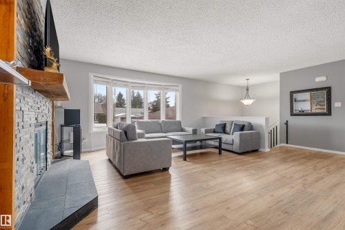 The living area features light-toned flooring, a stone-faced fireplace with a wooden mantel, and a bay window - 2437 89 Street, Edmonton, AB - Indoor Photo Showing Living Room With Fireplace