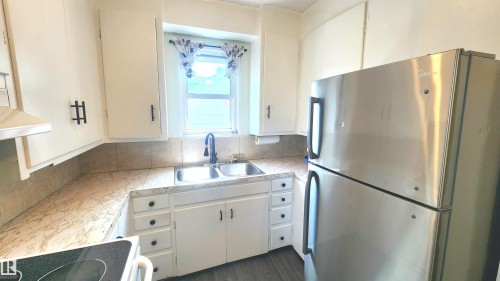 Kitchen featuring white cabinetry with dark hardware, a double basin sink with a gooseneck faucet, light-toned countertops, and a stainless steel refrigerator - 12139 81 Street, Edmonton, AB - Indoor Photo Showing Kitchen With Double Sink