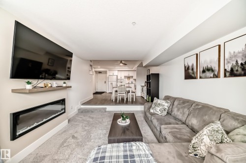This living space features light gray carpeting and a wall-mounted electric fireplace - 307 9504 182 Street Nw, Edmonton, AB - Indoor Photo Showing Living Room With Fireplace