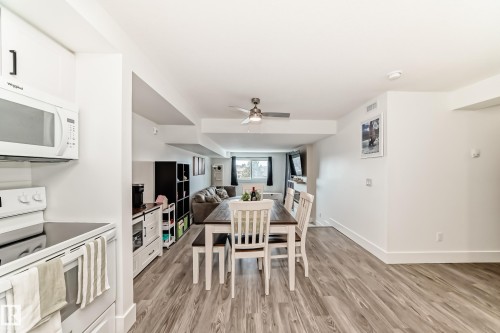 Open concept living area featuring wood-style flooring, a dining area with a wooden table and white chairs, and a kitchen area with white cabinetry and a white microwave oven - 307 9504 182 Street Nw, Edmonton, AB - Indoor