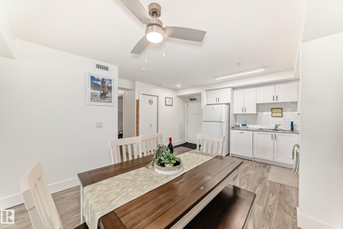 This open-concept area features wood-look flooring, a kitchen with white cabinetry and a white refrigerator, and a dining space with a wooden table - 307 9504 182 Street Nw, Edmonton, AB - Indoor