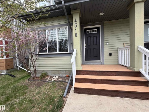 Exterior entry featuring a dark wood door with upper lites, light green horizontal siding, white window trim, wood-tone porch decking, and white railings - 4310 Prowse Link Link, Edmonton, AB - Outdoor