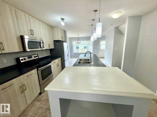 Kitchen featuring light wood-finish cabinetry, stainless steel appliances, dark countertops, and a central island with integrated double sink and pendant lighting - 4310 Prowse Link Link, Edmonton, AB - Indoor Photo Showing Kitchen With Double Sink With Upgraded Kitchen