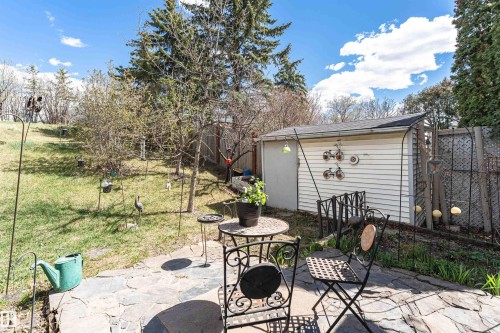 Stone patio area with wrought iron style outdoor seating, featuring a white vinyl-sided storage shed and a sloped grassy yard with mature trees - 1235 104 Street, Edmonton, AB - Outdoor With Deck Patio Veranda