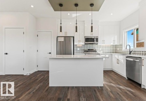 Modern kitchen featuring a central island with a light-toned countertop, stainless steel appliances, white cabinetry, and a patterned backsplash - 12440 47 Street, Edmonton, AB - Indoor Photo Showing Kitchen With Upgraded Kitchen