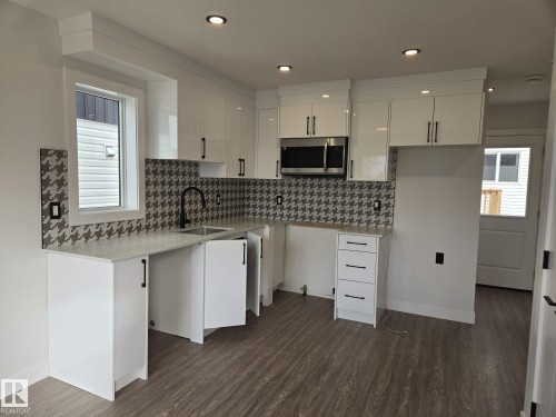 Kitchen featuring sleek white cabinetry, a patterned backsplash, stainless steel microwave, and wood-finish flooring - 12440 47 Street, Edmonton, AB - Indoor Photo Showing Kitchen With Upgraded Kitchen