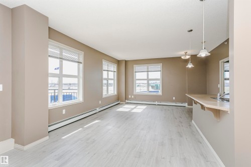 Open-concept living space featuring light wood-finish flooring, multiple large windows with blinds, a breakfast bar with an integrated sink, and overhead pendant lighting fixtures - 1-402 4245 139 Avenue, Edmonton, AB - Indoor Photo Showing Other Room