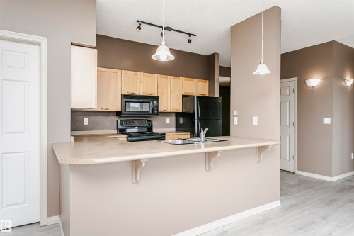 Kitchen featuring wood-finish cabinetry, black appliances, a double basin sink, and a breakfast bar with decorative corbels - 1-402 4245 139 Avenue, Edmonton, AB - Indoor Photo Showing Kitchen With Double Sink
