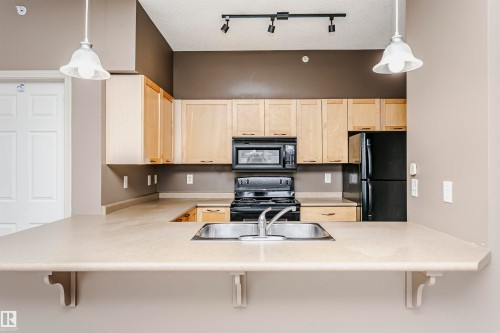 Kitchen featuring a breakfast bar with a double basin stainless steel sink, light wood cabinetry, built-in microwave, and black appliances - 1-402 4245 139 Avenue, Edmonton, AB - Indoor Photo Showing Kitchen