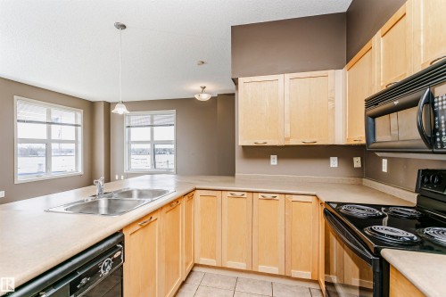 Kitchen featuring light wood-finish cabinetry, a double basin stainless steel sink, black built-in appliances, and light-toned countertops - 1-402 4245 139 Avenue, Edmonton, AB - Indoor Photo Showing Kitchen With Double Sink