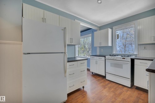 Kitchen featuring wood-finish flooring, white cabinetry, dark countertops, white appliance suite, and multiple windows - 4907 48 Avenue, Leduc, AB - Indoor Photo Showing Kitchen
