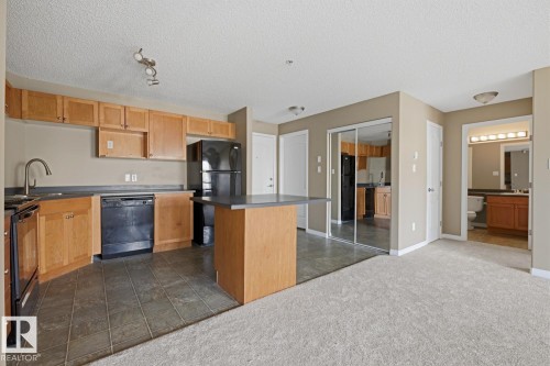 Kitchen and living area featuring wood cabinetry, black appliances, a central island with countertop seating, tiled flooring, and carpeted living space - 5106 7335 South Terwillegar Drive, Edmonton, AB - Indoor Photo Showing Kitchen