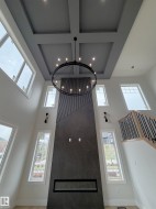 Two-story living area featuring a coffered ceiling, a large iron-finish chandelier, a floor-to-ceiling fireplace with an electric insert, tall windows, and a staircase with an iron-finish railing - 