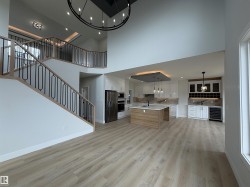 Two-story open concept living space featuring wood-finish flooring, a contemporary staircase with iron balusters, a kitchen island with a wood-finish base, high ceilings with an overhead chandelier, and a built-in wet bar with a beverage cooler - 