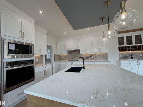 Spacious kitchen featuring a large island with a black sink, white cabinetry, integrated stainless steel appliances, marble-look countertops, and pendant lighting - 202 Edgewater Circle, Leduc, AB - Indoor Photo Showing Kitchen