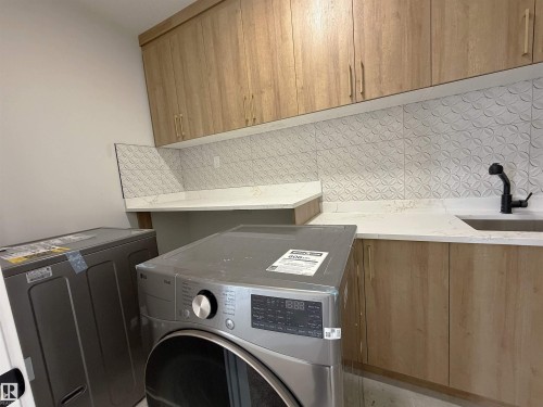 Laundry area featuring wood-finish cabinetry, white countertops, textured backsplash tiling, and a built-in sink with a matte black faucet - 202 Edgewater Circle, Leduc, AB - Indoor Photo Showing Laundry Room